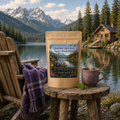 QuietMind Calm and Stress Support Botanical Blend by Oceanside Apothecary displayed on a rustic lakeside table beside a steaming mug, Adirondack chair with purple blanket, mountain lake, pine forest, and cabin.